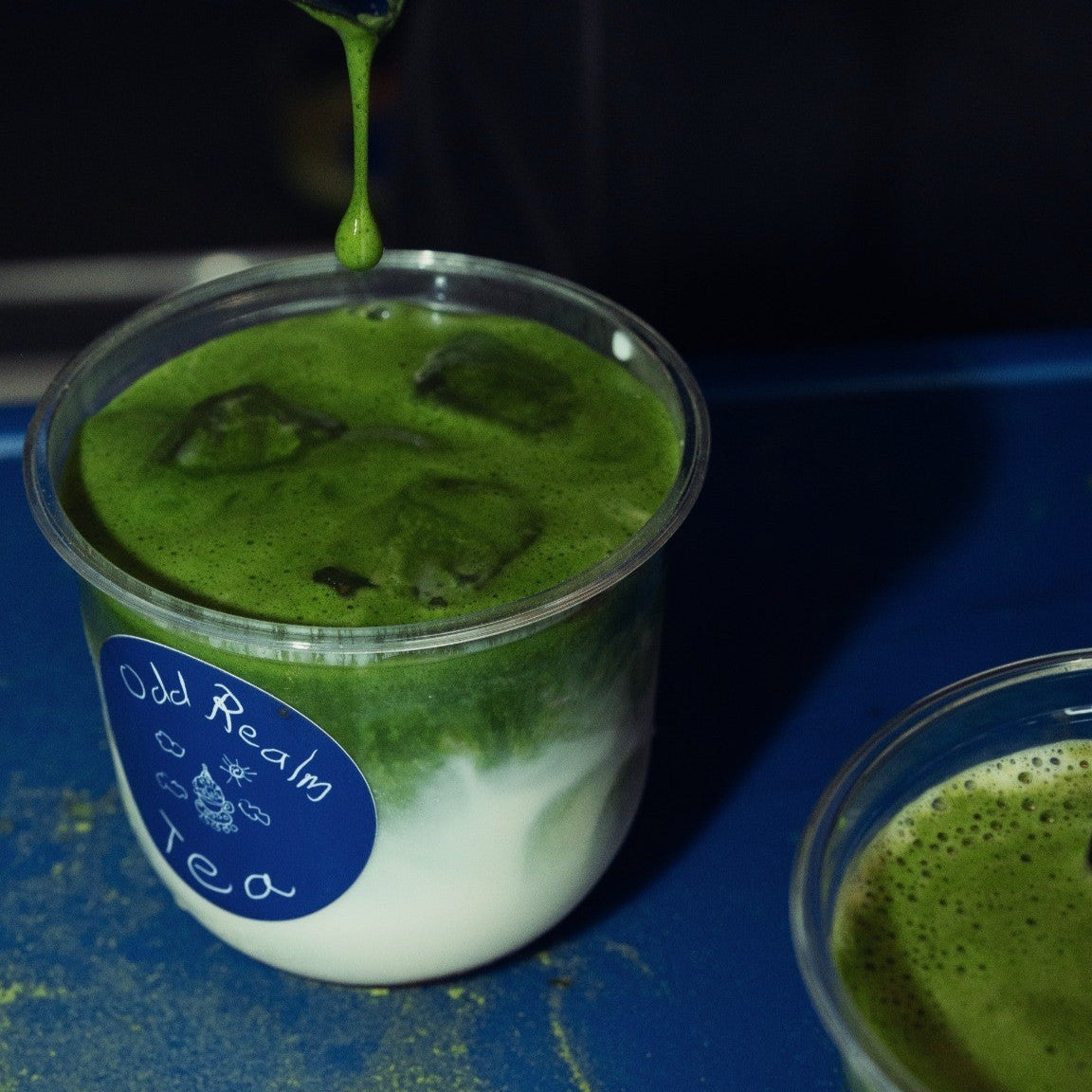 Eisgekühlter Matcha Latte mit OddRealm Blend Matcha No. 2 in Plastikbecher, serviert auf blauem Untergrund